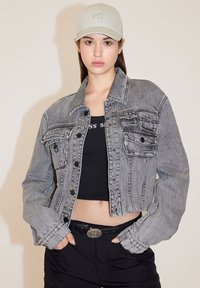 Chaqueta de mezclilla gris cortada con un cuello clásico y dos bolsillos en el pecho, combinada con una camiseta sin mangas negra y una gorra de color claro.