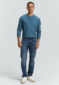 Camicia henley a maniche lunghe blu con tasca frontale, abbinata a jeans di lavaggio medio e sneaker bianche con dettagli beige.