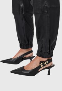 Zwarte leren spitse slingbackhakken met een gouden gesp, gedragen met zwarte elastische broek met boorden op een lichte achtergrond.