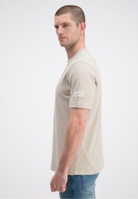 Beżowy t-shirt z krótkim rękawem, okrągłym dekoltem i małą metką na górnej części rękawa. Nosi się go z niebieskimi dżinsami, prezentowany z profilu.