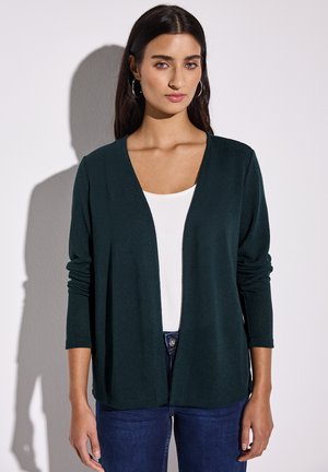 Mörkgrön, öppen cardigan i mjukt tyg med långa ärmar, kombinerad med en vit topp och blå jeans.