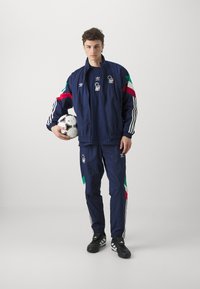 Námornícky atletický tréningový kabát a nohavice s červenými, zelenými a bielymi akcentmi. Obsahuje logo Adidas a náplety reprezentujúce Taliansko. Futbalová lopta držaná v ľavej ruke.