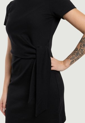 Frau in einem enganliegenden schwarzen Kleid mit seitlicher Bindedetail und einem floralen Tattoo am Unterarm, Hand auf der Hüfte.