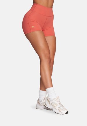 Zbliżenie osoby noszącej koralowe sportowe szorty z wysokim stanem oraz białe sneakersy New Balance i białe skarpety do kostek.