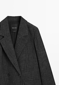 Tmavě šedý blazer s texturovaným tkaním, klopovými límci, dvouřadým zapínáním na knoflíky a jemným akcentovaným prošíváním. Matný povrch textilie.