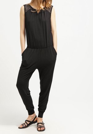 Sort ærmeløs jumpsuit med blonder ved skuldrene, tætsiddende ben, elastisk talje og sidelommer. Båret med flade sandaler.