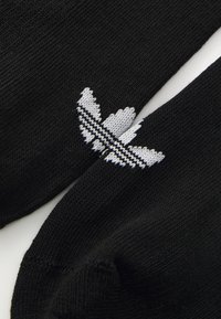 Juodos kojinės su raukšlėta tekstūra, kurių viršuje yra baltas siuvinėtas logotipas, primenantis sparnus, akcentuotas įstrižomis juodomis ir baltomis juostelėmis.
