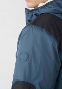 Giacca blu con dettagli neri, dotata di collo interno in pile e patch logo in gomma sulla manica. Tessuto liscio e design sportivo.