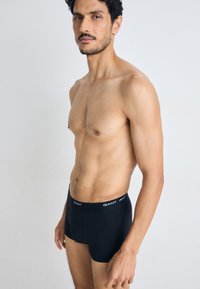 Boxeri negri dintr-un material neted, având o betelie elastică cu branding alb "GANT". Modelul stă cu o expresie neutră.