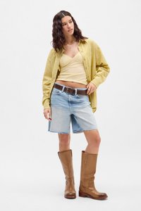 Chemise à carreaux jaune surdimensionnée, caraco beige, shorts en denim clair, bottes en cuir marron jusqu'au genou avec une finition texturée et une ceinture à boucle.
