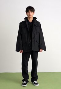 Jachetă neagră supradimensionată, cu buzunare mari în față, asociată cu pantaloni negri și pantofi negri. Textură netedă, design casual, elemente hardware simple.