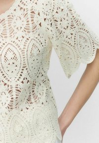 Blus i crema med spets, med intrikata blommönster, korta ärmar och vågiga kanter, i en lös passform och genomskinlig textur.