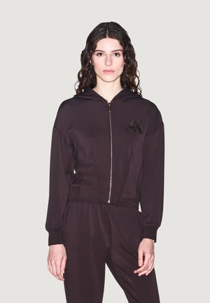 Femme aux longs cheveux bouclés portant un sweat à capuche noir zippé et un pantalon assorti, debout devant un fond clair uni.