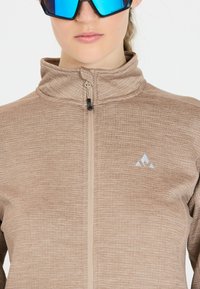 Lysebrune zip-up jakke med en struktureret overflade, høj krave og et blåt logo. Har en mellemlang lynlås og et slankt, moderne design.