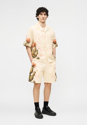 Young man with curly hair wearing beige short-sleeve shirt and matching shorts with cactus and flower prints, black socks, and black sneakers.