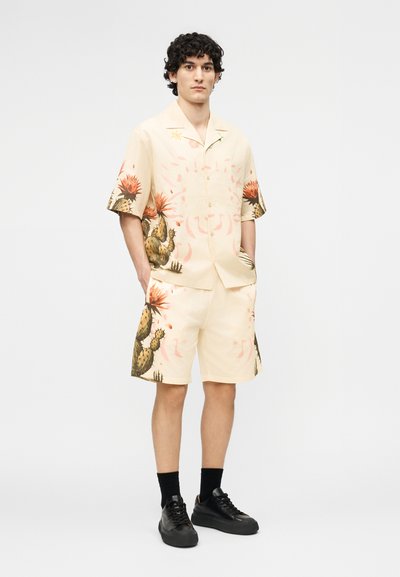 Young man with curly hair wearing beige short-sleeve shirt and matching shorts with cactus and flower prints, black socks, and black sneakers.