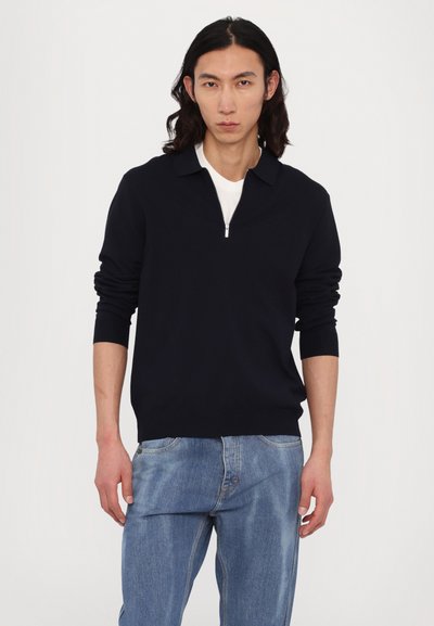 Tengerészkék cipzáras pulóver gallérral, hosszú ujjal és bordás mandzsettával, fehér ing felett viselve, világoskék farmernadrággal párosítva.