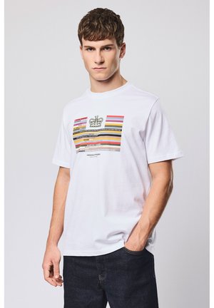 Junger Mann, der ein weißes T-Shirt mit mehrfarbigen horizontalen Streifen und Text trägt, kombiniert mit dunkler Jeans, posiert vor einem schlichten Hintergrund.