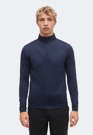 Giovane uomo con capelli biondi e ricci che indossa una maglia blu navy a maniche lunghe con zip e pantaloni neri, di fronte a uno sfondo uniforme.