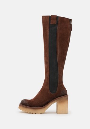 Botte en daim marron hauteur genou avec un panneau élastique noir, bout rond et un talon chunky beige. Comprend une fermeture éclair sur le côté pour un enfilage facile.
