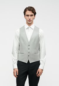 Noor mees, seljas helehall nööpidega vest, mis on valge särk ja mustade p�ükste kohal, seismas halli taustal.