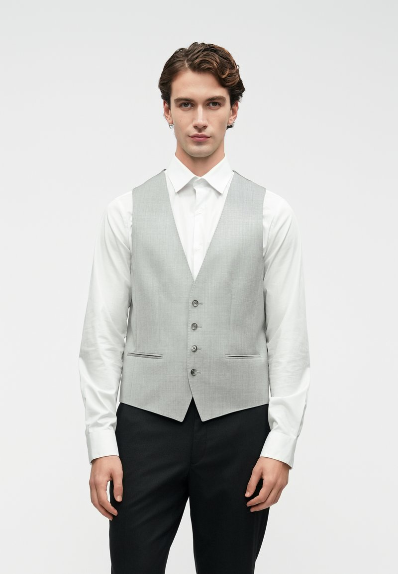 Noor mees, seljas helehall nööpidega vest, mis on valge särk ja mustade pükste kohal, seismas halli taustal.