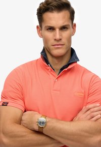 Polo en color coral con cuello de mezclilla y tapeta de tres botones. Presenta un logo en el pecho y se combina con un reloj en tono dorado.