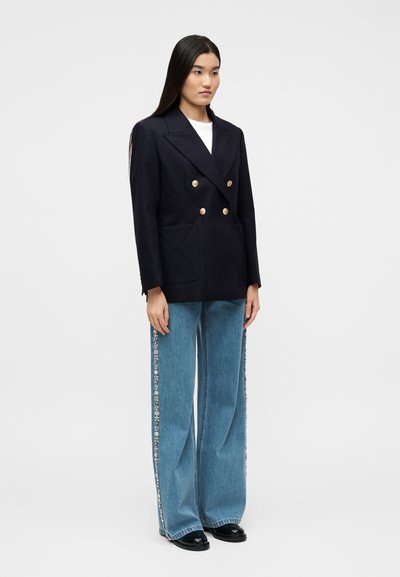Γυναίκα που φοράει μπλε σκούρο διπλό σακάκι με χρυσά κουμπιά, λευκή μπλούζα, φαρδιά μπλε τζιν με διακοσμητικά στο πλάι και μαύρα παπούτσια.