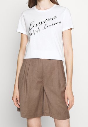 Kobieta w białej koszulce z krótkim rękawem z napisem „Lauren Ralph Lauren” oraz w brązowych spódnicowych szortach z wysokim stanem i plisami.