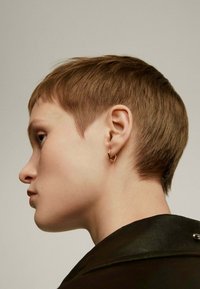 Pendiente de aro geométrico dorado usado con un peinado de cabello corto. El modelo tiene la piel suave y un fondo neutro. Lleva una camiseta oscura con detalle en el cuello.