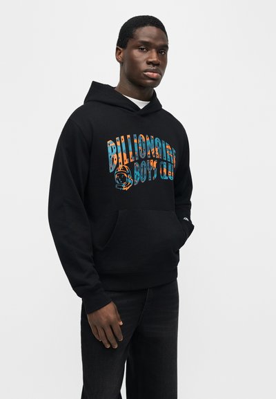 Άνδρας που φοράει μαύρη μπλούζα με κουκούλα με πολύχρωμο κείμενο "Billionaire Boys Club" και γραφικό με κράνος αστροναύτη, όρθιος με το ένα χέρι στην τσέπη.