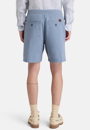 Uomo visto da dietro che indossa pantaloncini azzurri con tasche posteriori con bottoni, camicia a righe a maniche lunghe, calzini beige e scarpe da barca beige.