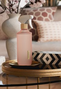 Lyserød ribbet sprayflaske med hvid aftrækker og gulddetalje, placeret på et rundt gyldent fad ovenpå en sort og hvid mønstret æske.