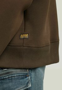 Bruine sweatshirt met geribbelde zoom en G-Star Originals Raw denim logo boven de jeanszak met zichtbare rits.