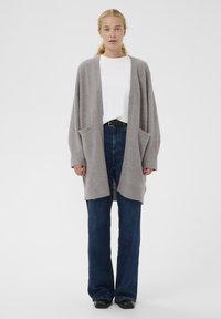 Cardigan grigio in maglia con apertura frontale, polsini a coste e due tasche frontali, abbinato a jeans a gamba larga blu e una maglietta bianca.