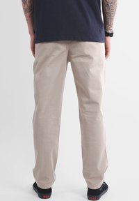 Pantalon en coton kaki au design droit, avec une texture lisse, associé à des chaussures noires. Poches arrière visibles et coutures apparentes.