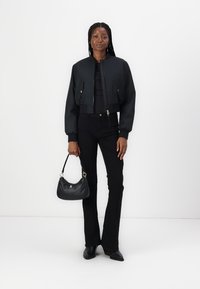 Zwarte cropped bomberjack met geribde boorden, gecombineerd met zwarte flarebroeken. Houdt een kleine zwarte handtas vast, staand tegen een witte achtergrond.