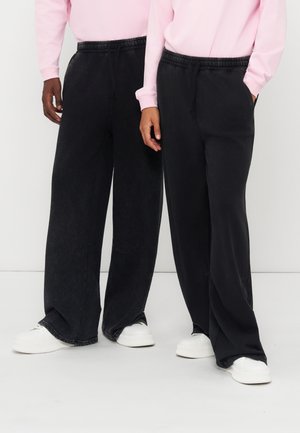 Deux personnes portant un pantalon noir à jambes larges, un haut rose clair à manches longues et des baskets blanches, debout côte à côte.