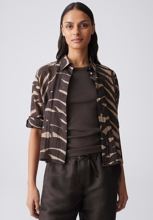Femme portant une chemise boutonnée à motif zèbre sur un haut côtelé foncé et un pantalon ample à cordon, debout devant un fond clair uni.