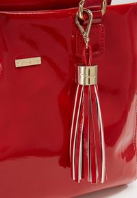 Borsa a mano rossa lucida con finiture in oro, caratterizzata da una frangia decorativa con fili rossi e bianchi. Piccola etichetta del marchio visibile all'esterno.