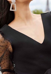 Black V-neck dress with textured fabric and sheer polka dot sleeves. Large diamond-shaped earrings with rhinestones.