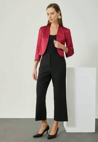Red satin jacket over a black jumpsuit, featuring cropped trousers and a fitted silhouette. Model wears black heels and gold earrings.