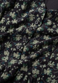 Zwarte stof met groene bloemmotieven en blauwe accenten. Voorzien van zwarte knopen en een gestructureerd corduroy oppervlak. Uitlopende ontwerp.
