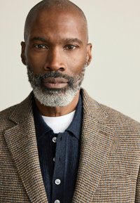Brown houndstooth blazer with textured fabric, worn over a navy button-up shirt. Features a white undershirt and prominent facial hair.
