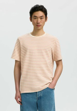Ung mand med sort hår iført en beige og orange stribet kortærmet skjorte og blå jeans, stående med den ene hånd i lommen.