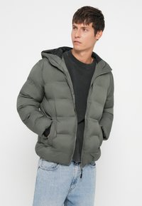 Πετσέτα μπουφάν σε ελιά χρώμα, με κουκούλα, φερμουάρ και δύο πλαϊνές τσέπες. Φορεμένο πάνω από μαύρο πουκάμισο και ανοιχτό γαλάζιο τζιν.