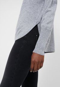 Pull en maille grise avec des poignets côtelés, design superposé sur le côté. Associé à un jeans noir avec un petit accent doré sur la poche.