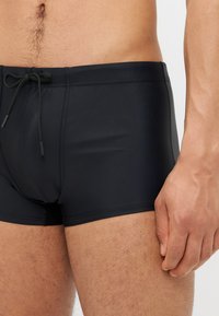 Shorts de bain noirs avec une taille à cordon de serrage, présentant un matériau lisse et extensible ainsi qu'une coupe ajustée, mettant en valeur une silhouette élancée.