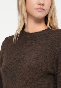 Donna bionda che indossa un maglione di lana scura e morbida, mostrata dalla nuca fino a metà torace su uno sfondo bianco uniforme.