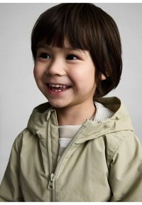 Giacca beige con cappuccio, chiusura a zip, texture visibile e design leggero. Sotto indossa una maglietta a righe, ha i capelli corti e un'espressione sorridente.